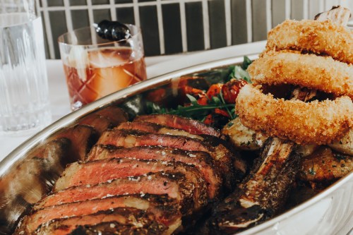 A platter of sliced steak, crispy onion rings, and roasted vegetables, accompanied by a cocktail in the background.