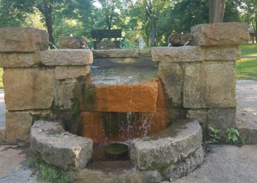 A stone fountain with water flowing over rocks, surrounded by greenery in a park setting.