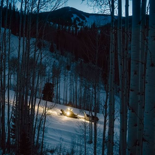A snowcat vehicle lights up a snowy slope at dusk, surrounded by tall trees and mountains in the background.