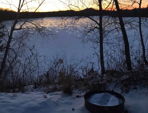 A serene winter scene of a frozen lake at sunset, framed by bare trees and snow-covered ground.