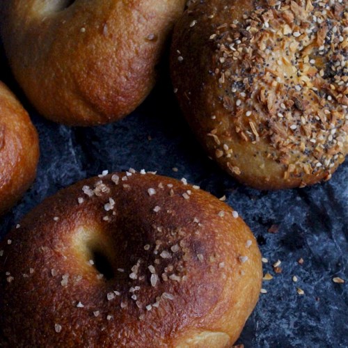 A close-up of four bagels, two topped with seasoning and two with coarse salt, on a dark surface.
