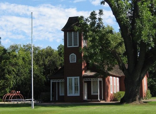 A red wooden building with a clock tower, surrounded by green grass and trees, under a blue sky with clouds.