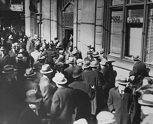 A crowded street scene from the 1930s, with many people in hats gathered outside a building.