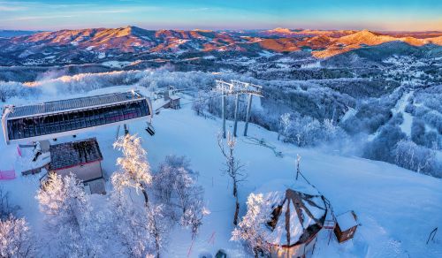 A snowy mountain landscape at sunrise, featuring ski lifts and frosted trees with distant hills in the background.