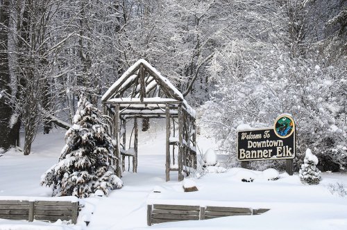 A snowy scene featuring a welcome sign for Downtown Banner Elk, surrounded by snow-covered trees and a rustic structure.