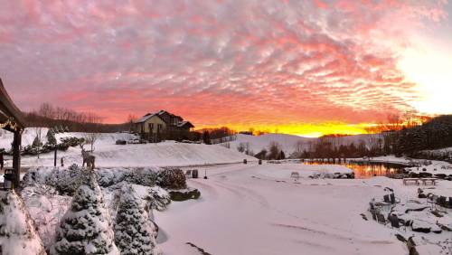 A snowy landscape at sunset, featuring a house, trees, and a serene lake under a colorful sky.