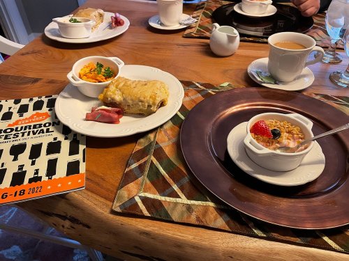 A table setting with a plate of breakfast casserole, a fruit dish, and a bowl of yogurt with cereal, alongside a festival brochure.