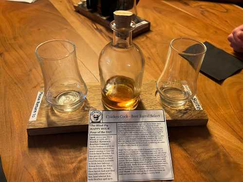 A wooden tray holds a bottle of Chicken Cock whiskey and two empty glasses, with a menu card detailing the drink.