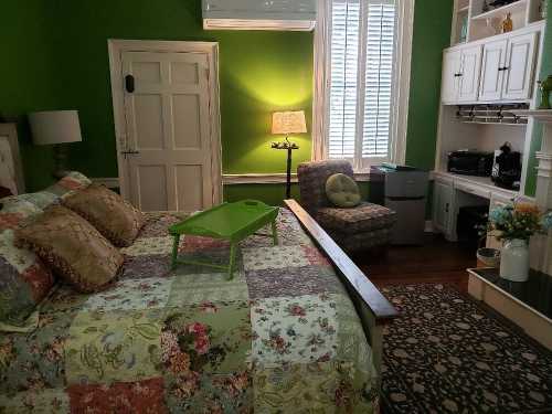 Cozy bedroom with green walls, a quilted bed, a small table, a lamp, and a comfortable chair in the corner.