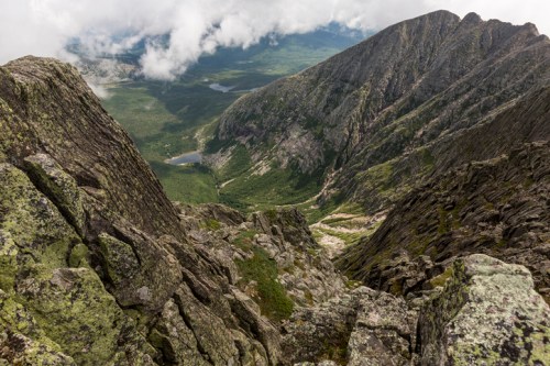A breathtaking view from a mountain peak, showcasing rocky cliffs and a lush valley below, with clouds overhead.