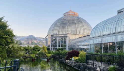 A large glass conservatory with domed roofs, surrounded by lush greenery and a tranquil pond.