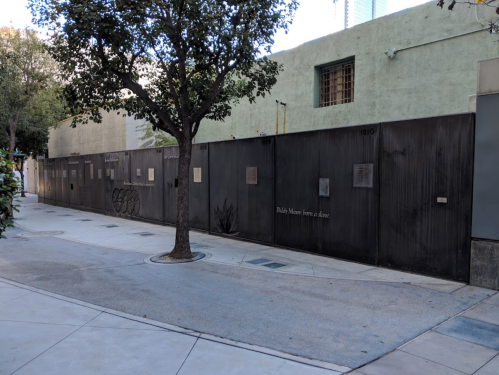 A metal fence along a sidewalk, with trees and buildings in the background, featuring plaques and text on the fence.