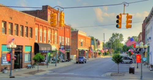 A quiet small-town street lined with brick buildings, shops, and American flags, under a clear blue sky.