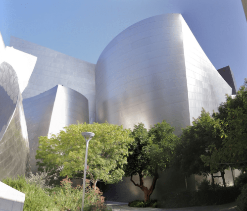 A modern building with curved metallic surfaces, surrounded by greenery and trees under a clear blue sky.