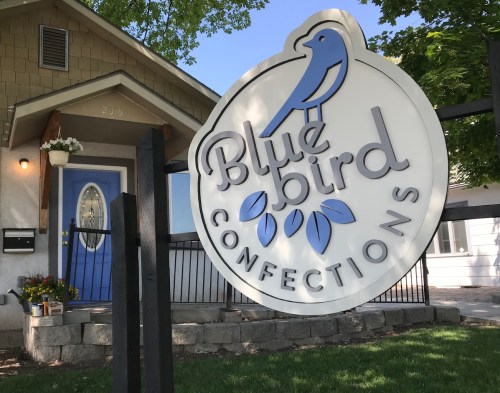 Sign for "Blue Bird Confections" in front of a house, featuring a blue bird logo and a welcoming entrance.