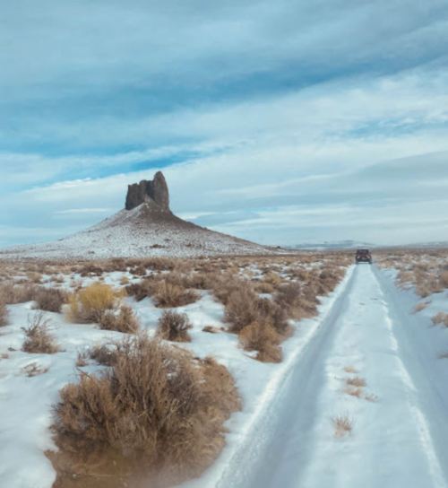 A snowy landscape with a dirt road leading to a distant rock formation under a cloudy sky.