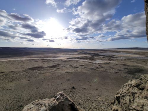 A vast, open landscape under a cloudy sky, with the sun shining in the background and rocky terrain in the foreground.