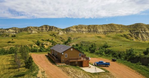 A brown house with a blue truck parked nearby, set against rolling green hills and a clear blue sky.