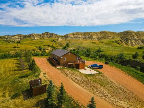 Aerial view of a brown house on a dirt road, surrounded by green hills and trees under a partly cloudy sky.