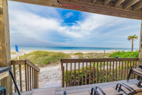 View of a sandy beach and ocean from a wooden deck, with palm trees and a cloudy sky in the background.