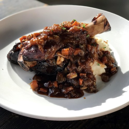 A plated dish featuring a braised meat shank topped with a rich sauce and served over creamy mashed potatoes.