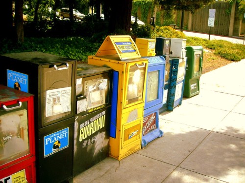 A row of colorful newspaper vending machines on a sidewalk, surrounded by greenery and trees.
