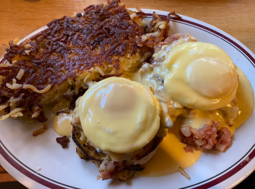 A plate of eggs Benedict with ham and hollandaise sauce, served with crispy hash browns on the side.