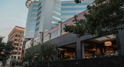 A modern building with large windows, featuring a restaurant patio and trees in the foreground, set against a city skyline.