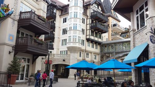 A charming village square with European-style buildings, outdoor seating, and blue umbrellas on a sunny day.