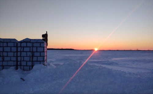 A snowy landscape at sunset, with a silhouette of a structure in the foreground and vibrant colors in the sky.