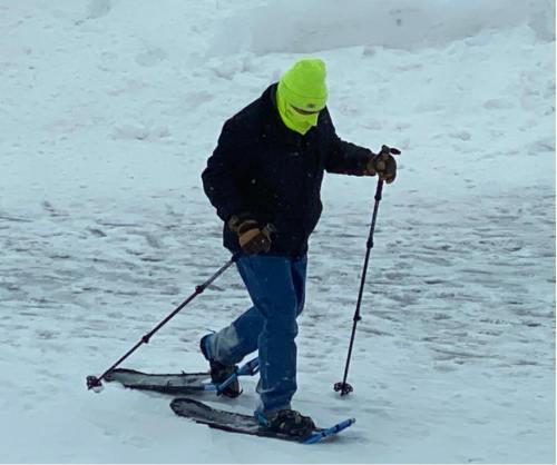 A person in a bright yellow hat and mask snowshoes through a snowy landscape, using trekking poles for support.