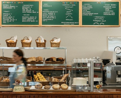 A café counter displaying a menu, baked goods, and a barista in motion, with baskets of bread and pastries.