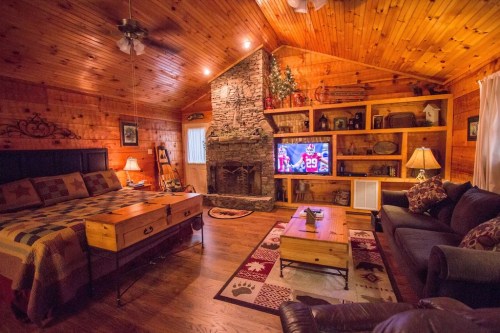Cozy cabin interior with wooden walls, a stone fireplace, a TV, and a bed with a quilt, surrounded by rustic decor.