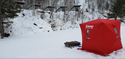 A bright red ice fishing shelter on a snowy landscape, with cabins visible in the background among trees.