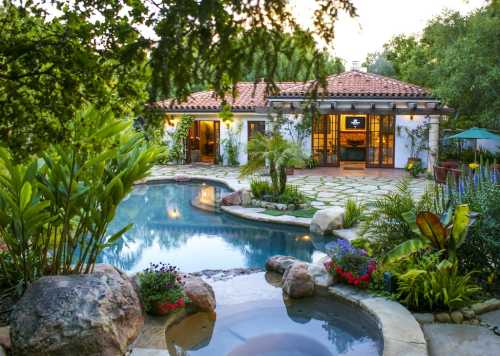 A serene backyard with a pool, lush greenery, and a charming house with a tiled roof, surrounded by colorful flowers.