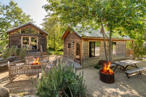 Two rustic cabins surrounded by trees, with a fire pit and seating area in a serene outdoor setting.