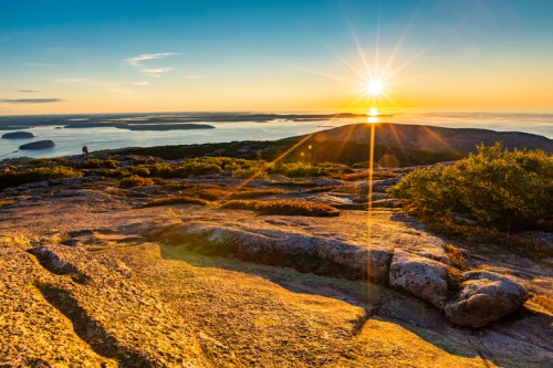 Sunrise over a rocky landscape with distant islands and a shimmering sea, casting a warm golden glow.