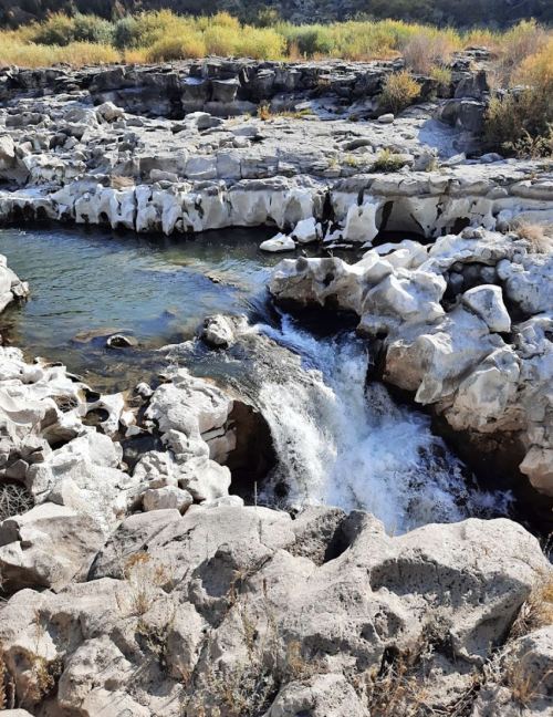 A serene waterfall cascades over rocky terrain, surrounded by lush greenery and rugged stone formations.