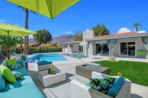 A sunny backyard with a pool, lounge area, and green umbrellas, surrounded by palm trees and mountains in the background.