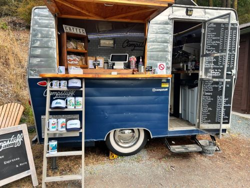 A vintage blue food truck with a wooden counter, serving coffee and snacks, surrounded by nature.