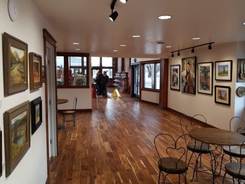 A spacious art gallery with wooden floors, featuring framed paintings on the walls and two people in the background.