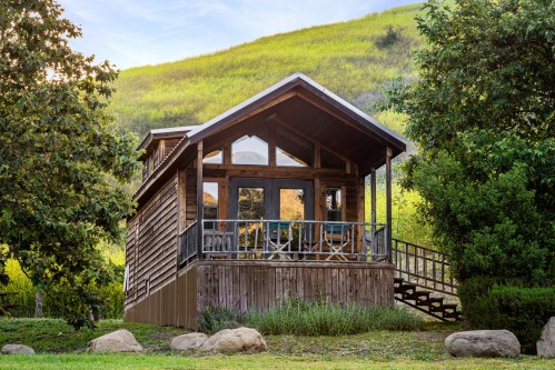 A cozy wooden cabin with a porch, surrounded by greenery and hills under a clear sky.