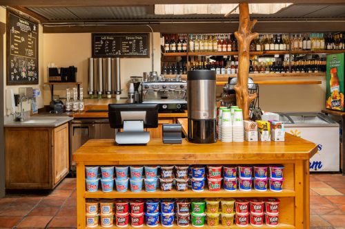 A cozy café counter with a coffee machine, drinks, and shelves of snacks and ice cream tubs.