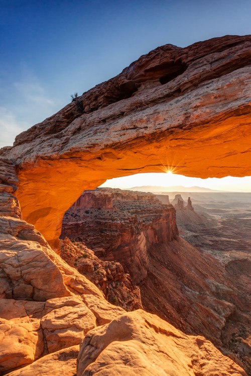 A stunning rock arch frames a sunset over a vast canyon landscape, highlighting warm orange and earthy tones.