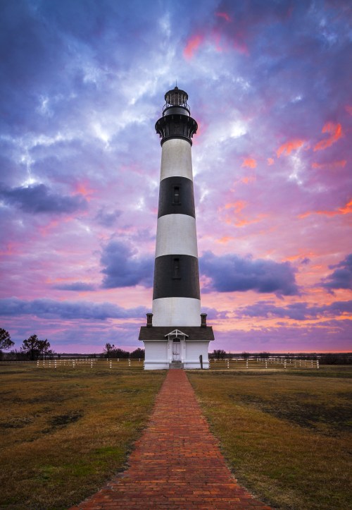 A tall black and white lighthouse stands against a colorful sunset sky, with a path leading to its entrance.