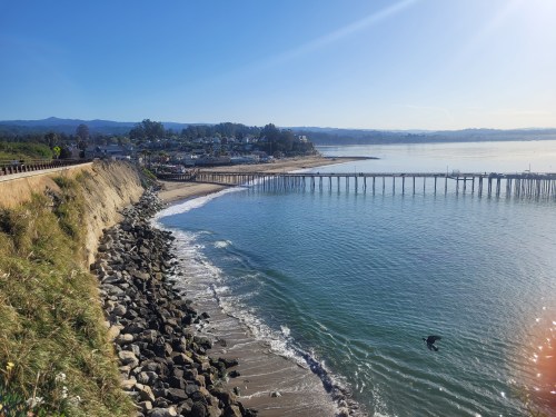 A scenic view of a calm beach with a pier, rocky shoreline, and clear blue sky in the background.