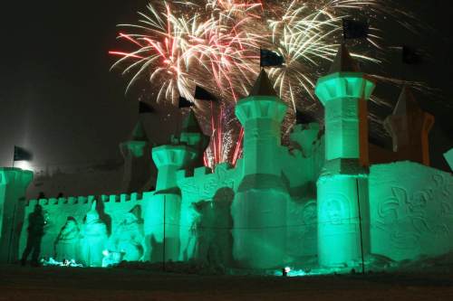 A snow castle illuminated in green with fireworks bursting in the night sky above it.