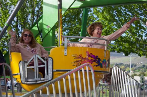 Two women in matching sweatshirts wave from a yellow carnival ride, surrounded by green trees and a festive atmosphere.