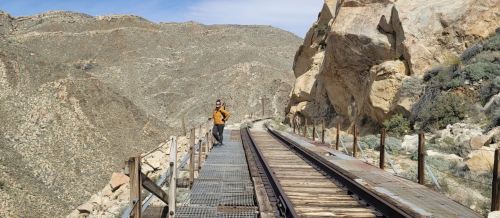 A person stands on a narrow railway bridge surrounded by rocky mountains and a clear blue sky.