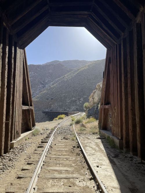 View from inside a wooden tunnel, looking out at a winding railway and mountainous landscape under a clear sky.